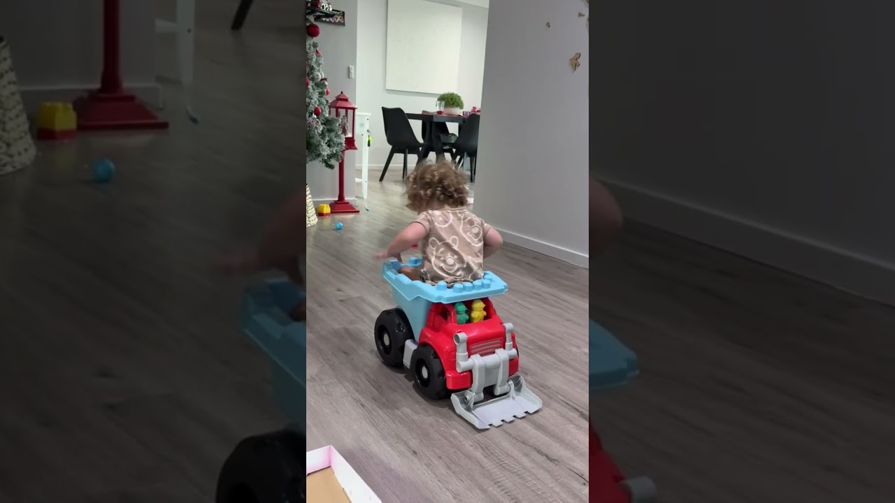 Little boy sits in back of toy dump truck then falls forwards and faceplants near Christmas tree