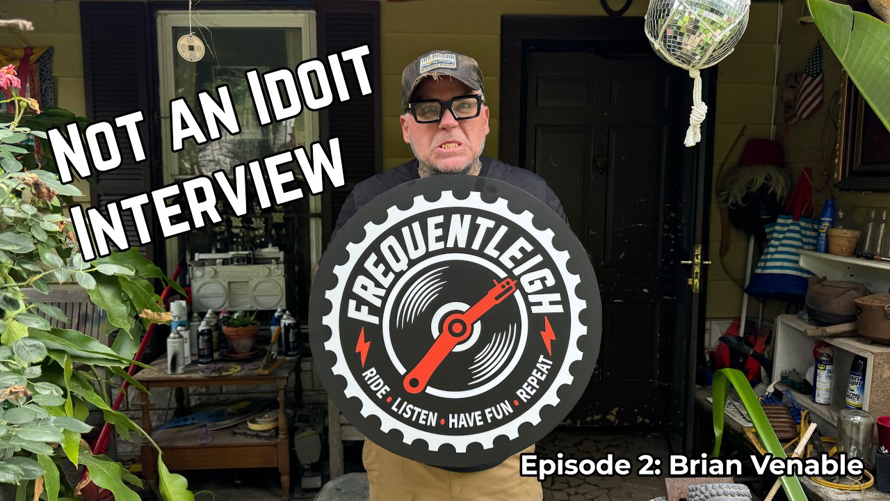 Brian Venable of Lucero grimaces on his front porch holding a round FrequentLeigh chainring logo sign. Text reads "Not an Idiot Interview, Episode 2: Brian Venable.