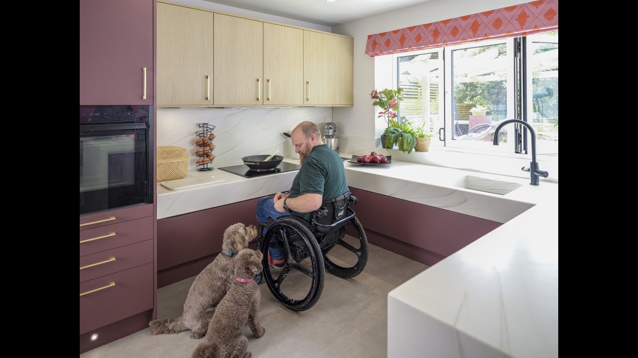 Video shows a wheelchair user in his wheelchair, in a large accessible kitchen, with two dogs at his side looking up at him.