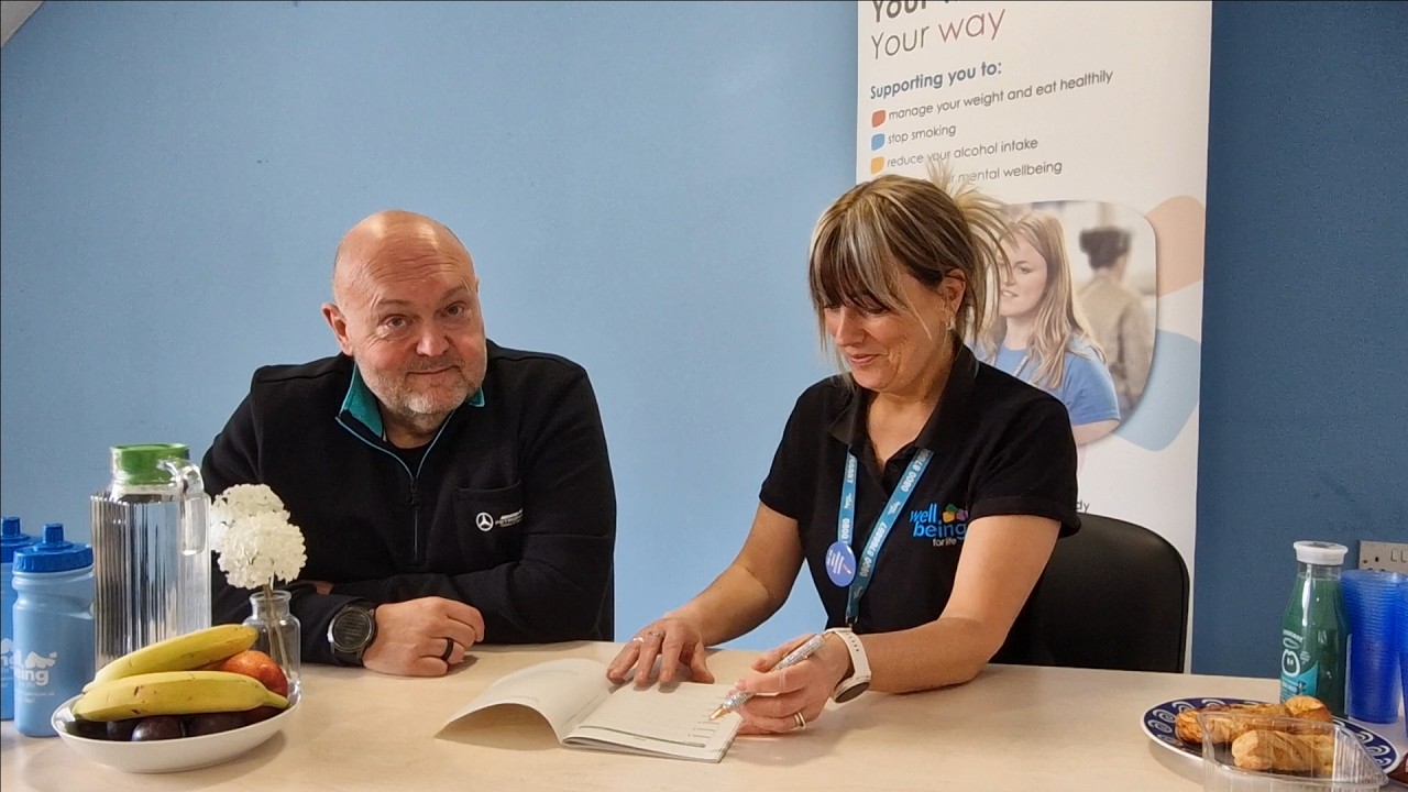 Chris sits with Sharon at a table. There is fruit and water bottles on the table and they are working through an Eatwise session booklet.