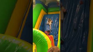 Girl climbs up wall of bounce house slide then falls then lands on kid