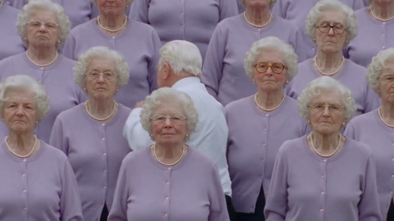 Storm II remade as a grandma choir: rows of elderly women in lilac cardigans and pearls around a white-haired man in a white shirt