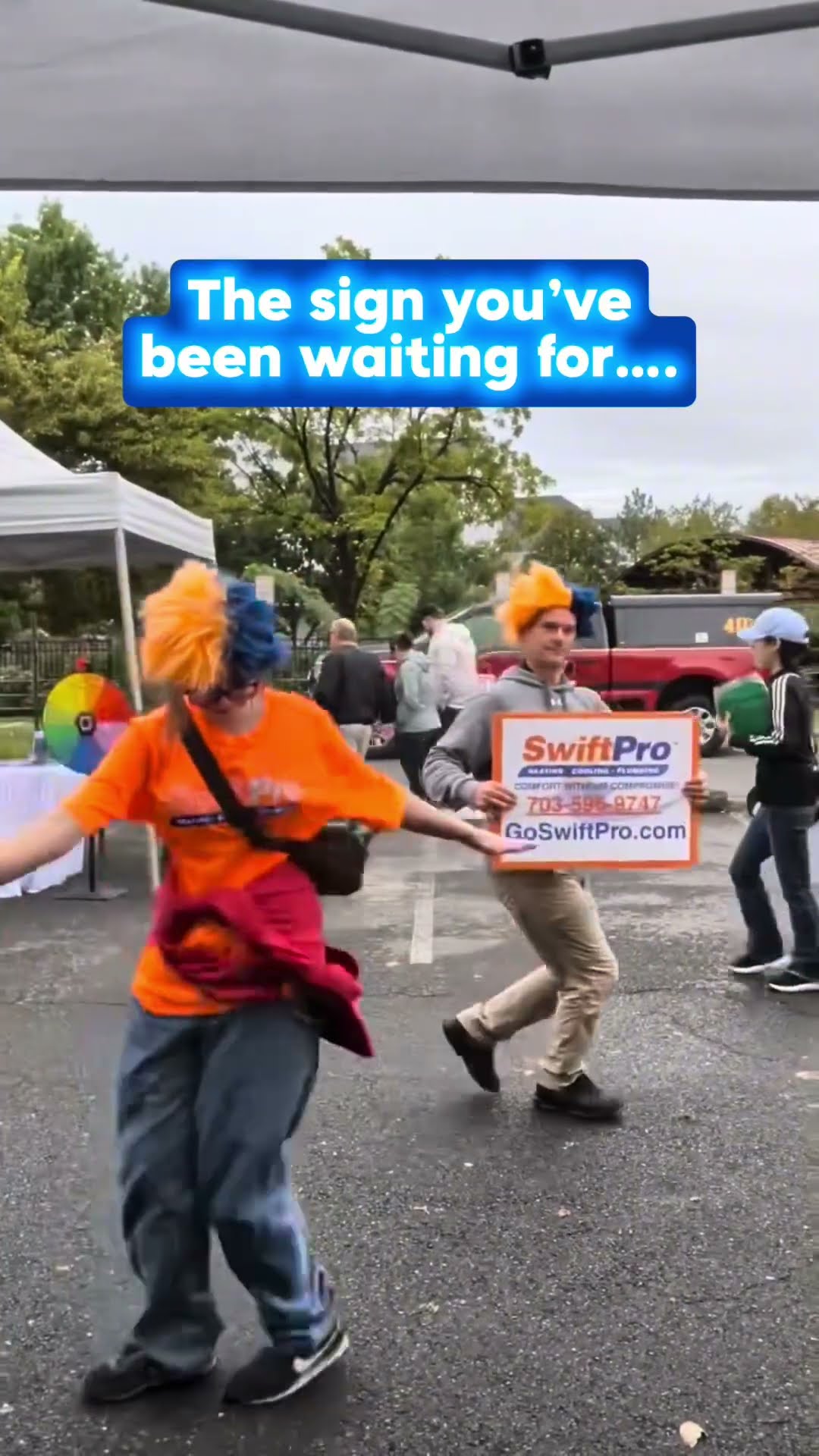 Outdoor community event under tents: a person in an orange SwiftPro t-shirt and orange-and-blue wig dances while a man in a grey hoodie dances holding a SwiftPro yard sign with the phone number and Go