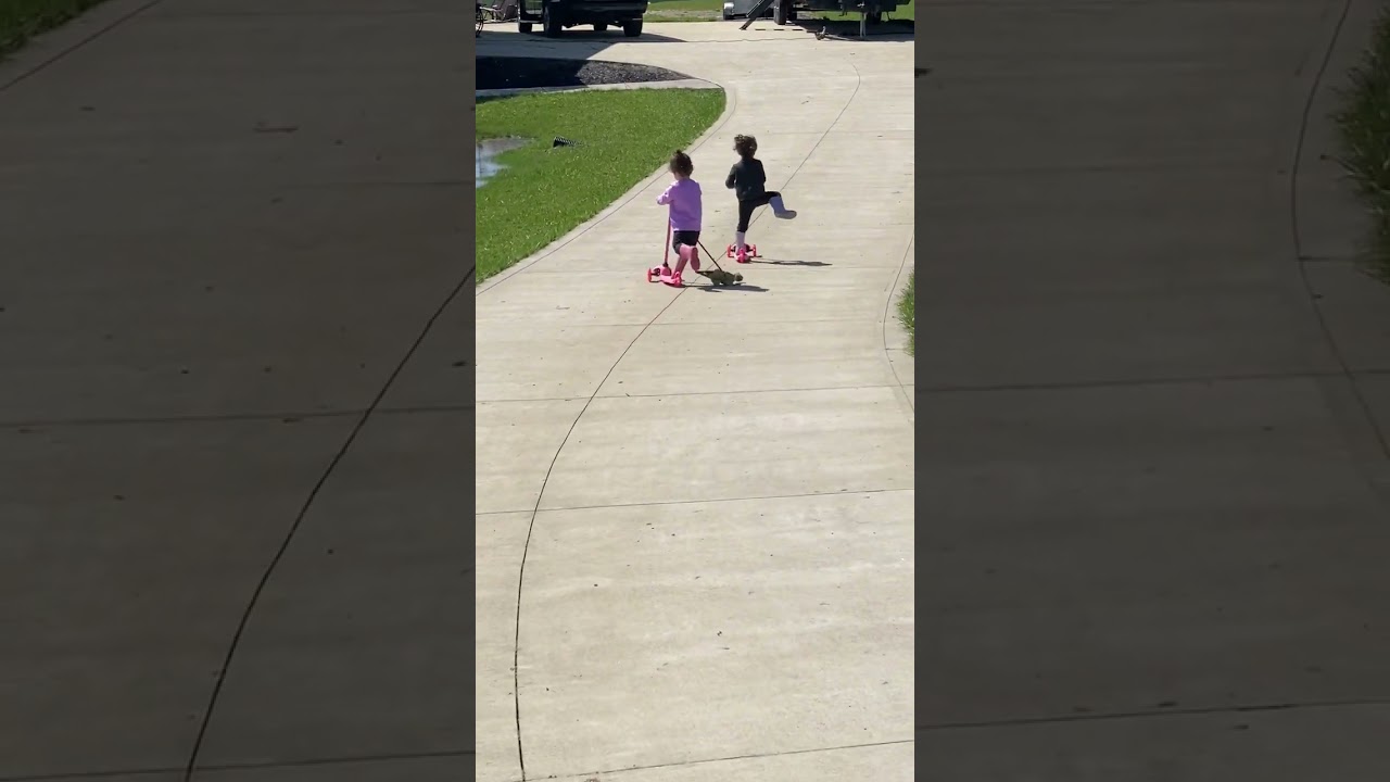 Two little girls ride scooters down driveway and they both fall one after the other