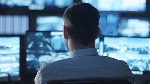Security Officer is Working on Computer in a Dark Monitoring Room Filled with Display Screens |