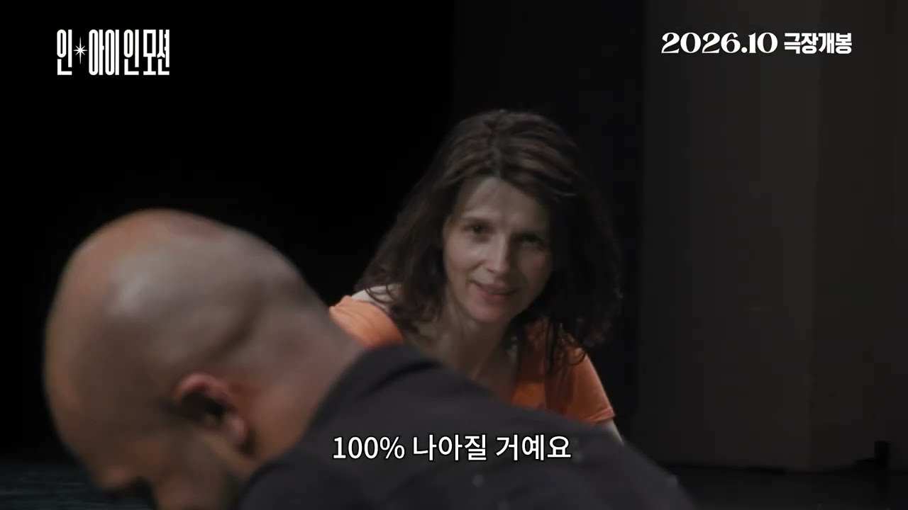 어두운 무대에서 몸을 숙인 민머리 남성과 그 뒤의 곱슬머리 여성