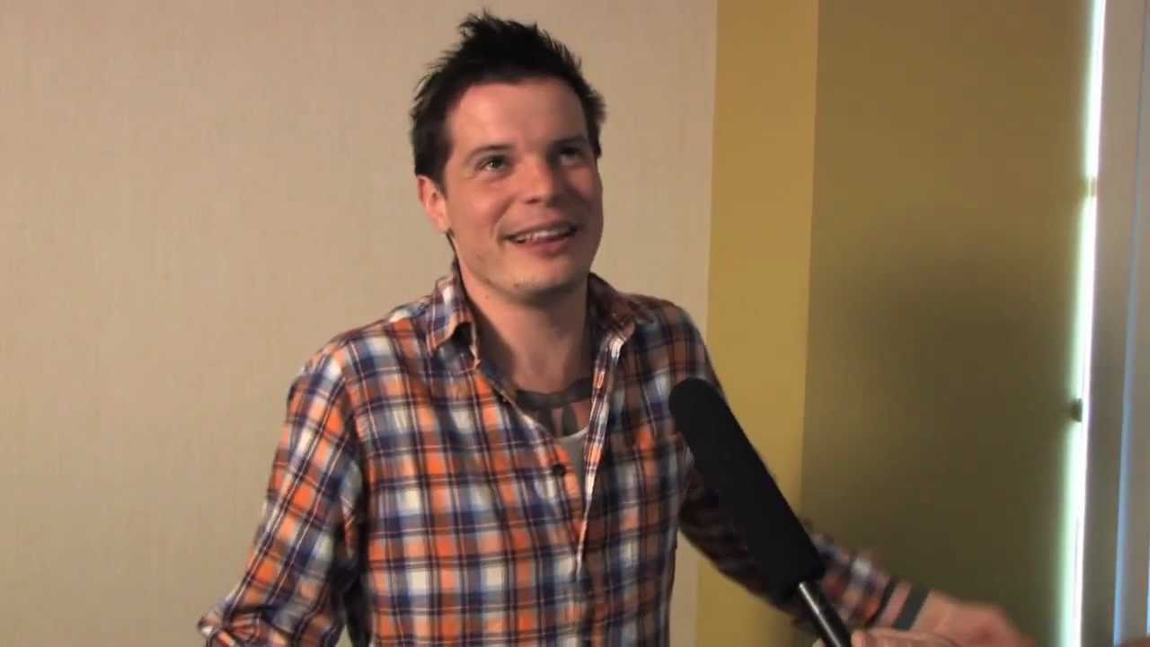 A screenshot from an interview with BackstageWithBrian on YouTube. A man, James Zinkland, sits in a chair being interviewed online. He wears a checkered shirt and has dark, spikey hair. 