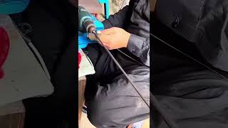 Smoothing a long metal rod with sandpaper using an electric hand drill at a workshop table