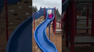 Little girl in pink rain coat and boots rides down blue slide then lands in a center split