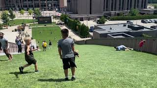 Woman rolls down grass hill and over little boy in yellow shirt