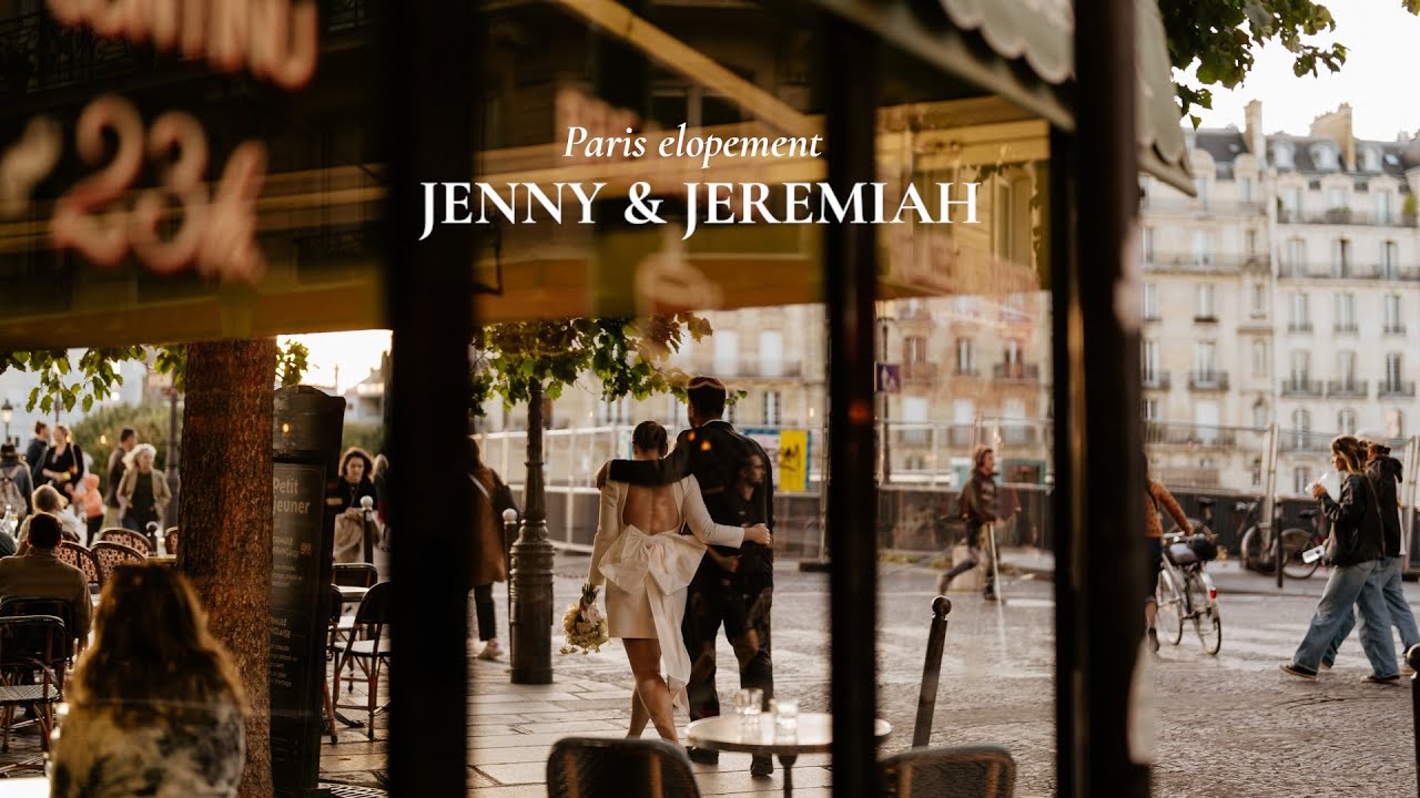 Bride and groom walk arm in arm along a Paris street in warm light, seen through a café window with guests nearby