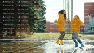 Happy girls friends jumping dancing in puddle wearing raincoats and rainboots