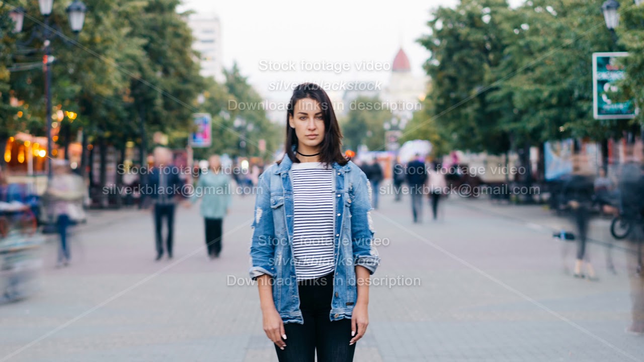 Time-lapse of unhappy lonely girl standing alone in the street and looking at camera when people are