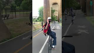 Guy waves and attempts to sit on middlebar of bike while holding skateboard on backpack