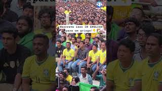 A huge crowd of people sits in a public space, and they look at a screen.