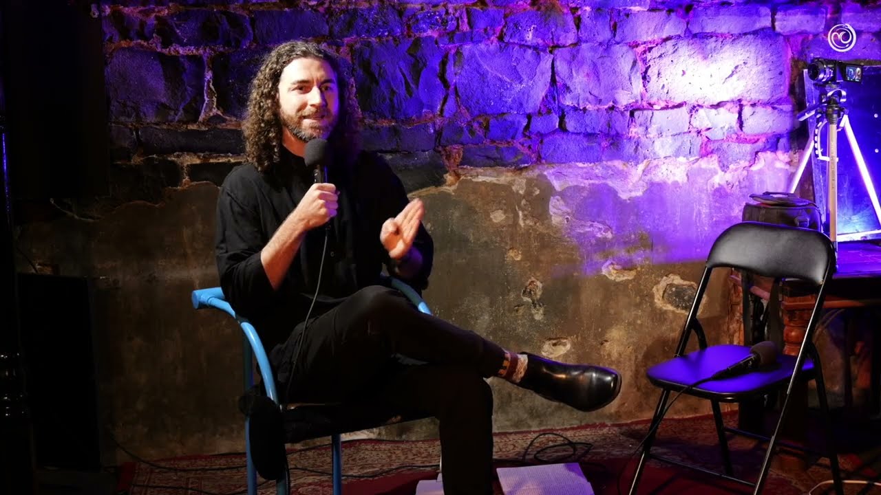 Tim Adalin seated on a blue chair with a microphone, speaking in a brick vault washed in lavender light, at Underground Philosophy.