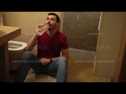 Young bearded drunk man is sitting near the toilet holding his belly and drinking pure water