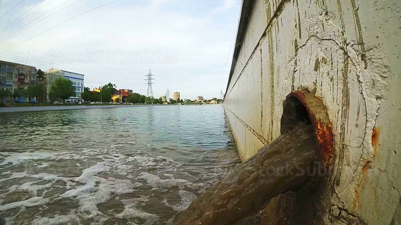 A rusty storm drain pipe with storm water runoff flowing out of it into a river. The drain carries