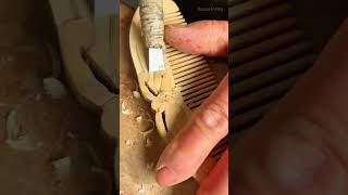 Carving a light brown wooden comb precisely using a sharp metal chisel tool in a bright indoor