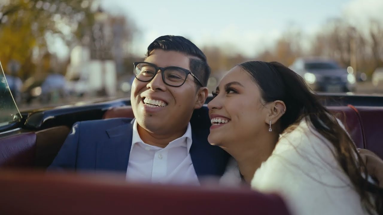 A smiling bride and groom sit close together in an open car on a sunlit Paris street