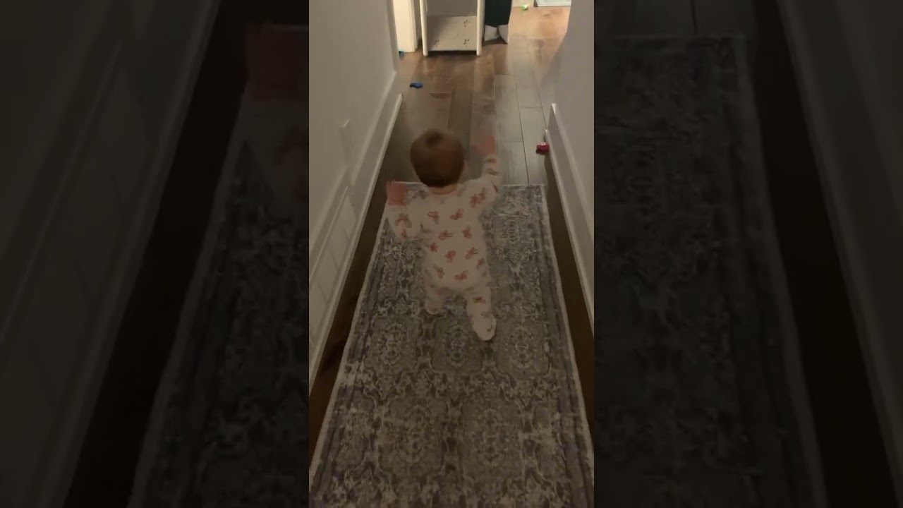 Baby girl in onesie pajamas takes first steps through hallway then sister gives a hug and both fall
