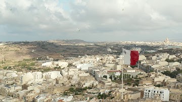 Flag of Malta waving in Wind with view of City on Gozo Island, Aerial Close up slide left
