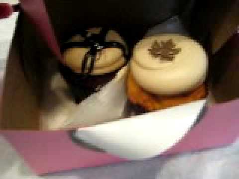 Witness the unboxing of two of the first cupcakes sold at the new Georgetown Cupcake at Bethesda Row, in downtown Bethesda, Maryland. They were purchased at the Grand Opening on Bethesda Avenue. On the left is a Peanut Butter Fudge cupcake, and a Pumpkin Spice is on the right. The Peanut Butter Fudge has fudge sauce in the center, like a lava cake. The Pumpkin Spice has a large quantity of maple frosting, and a chocolate maple leaf decoration with a special effect that gives the edible leaf a metallic appearance.