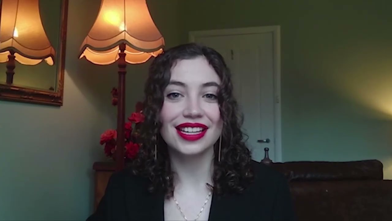 Amy Cripps smiling during a video interview, sitting indoors with a lamp and flowers in the background.