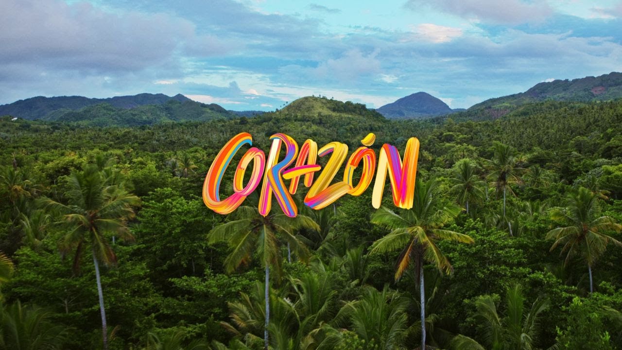 Vista aérea de una selva tropical con palmeras y montañas al fondo, con la palabra 'CORAZÓN' escrita de forma colorida y artística en el centro de la imagen.