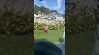 Little boy in red bathing suit sprays himself on his face with garden hose while dog watches