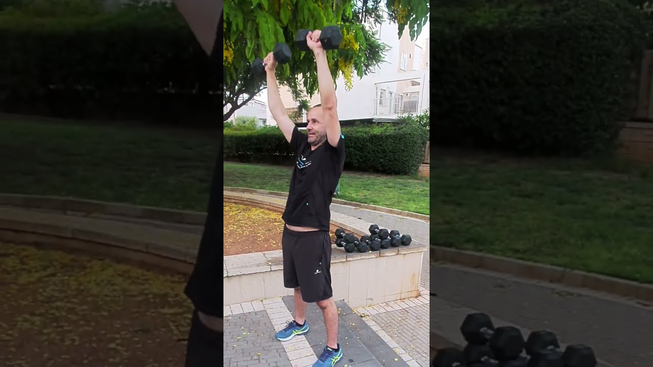 מתאמן מרים משקולת יד מעל הראש באימון ערב בפארק, משקולות מסודרות על הרצפה לצידו