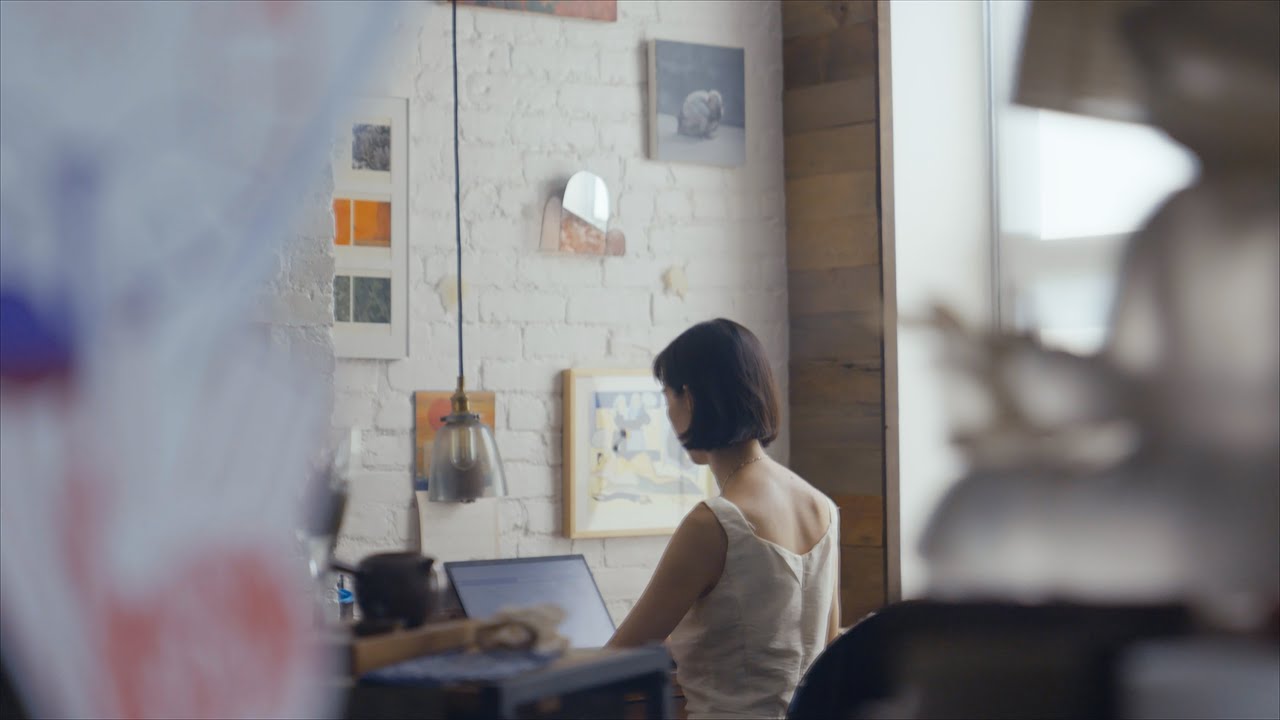 Color still from The Story We Tell (dir. Jackie Yunchang Zhang, 2025). A woman with short hair is seen in a mirror reflection, sitting at a desk facing a white brick wall, working on a laptop. 