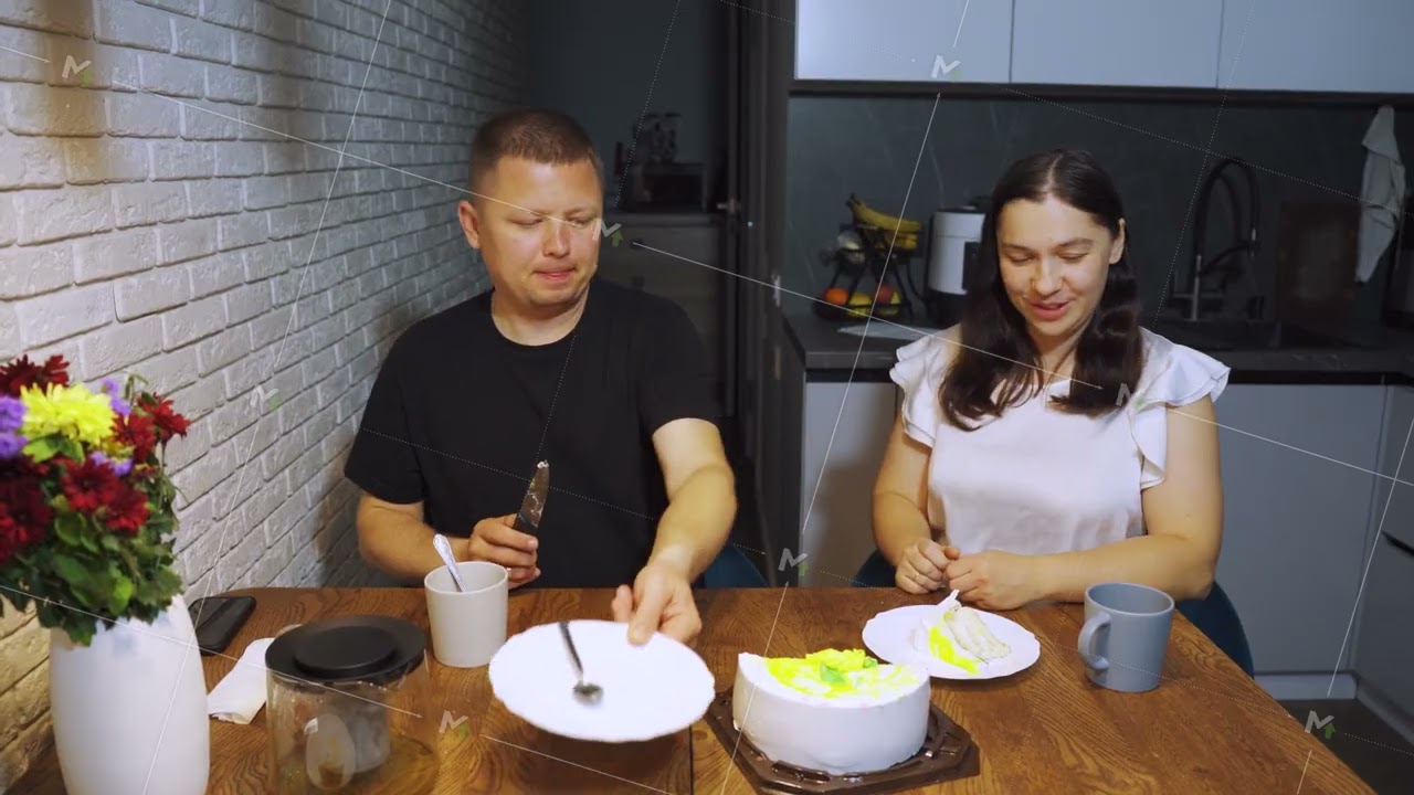 A man cuts a cake at home in the kitchen, sitting at the dining table with his wife. A modest