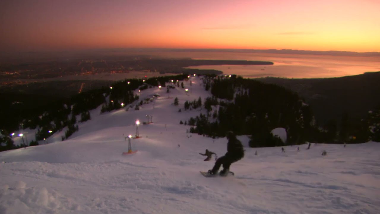 Static shot of various snowboarders travelling down Grouse Mountain with Vancouver in background