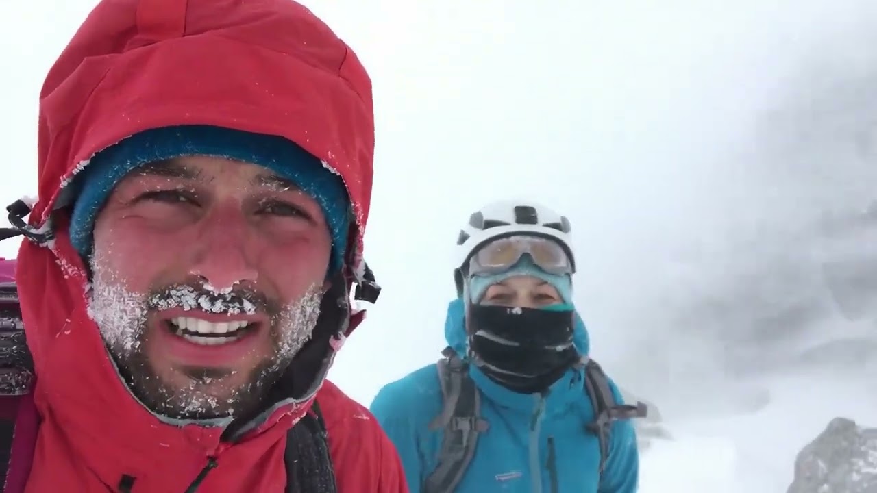 1550 Two mountain explorers in bright technical shells, ice crusted faces, eyes narrowed wi