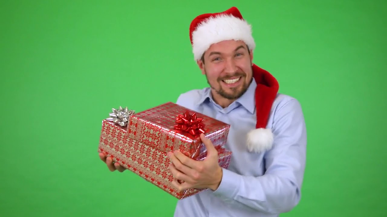 A young handsome man in a Christmas hat holding a present dances with a smile green screen studio