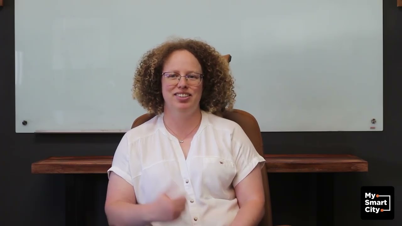 Woman with curly hair and glasses sitting in front of a white board, Nicola, product manager of My Smart City