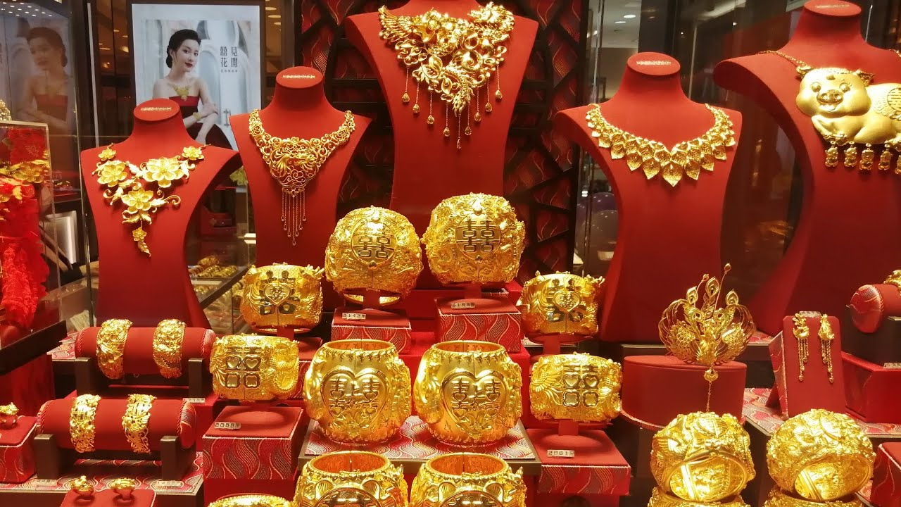 A display of elaborate gold jewelry, including necklaces, bracelets, rings, and ornamental accessories, is showcased on red busts and stands in a Hong Kong jewelry shop, with some pieces featuring traditional Chinese symbols and motifs.