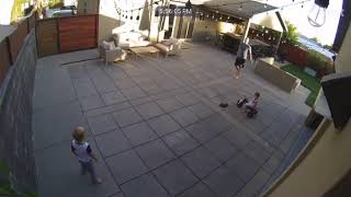 Guy throws football at wall that bounces and hits kid seated on tricycle