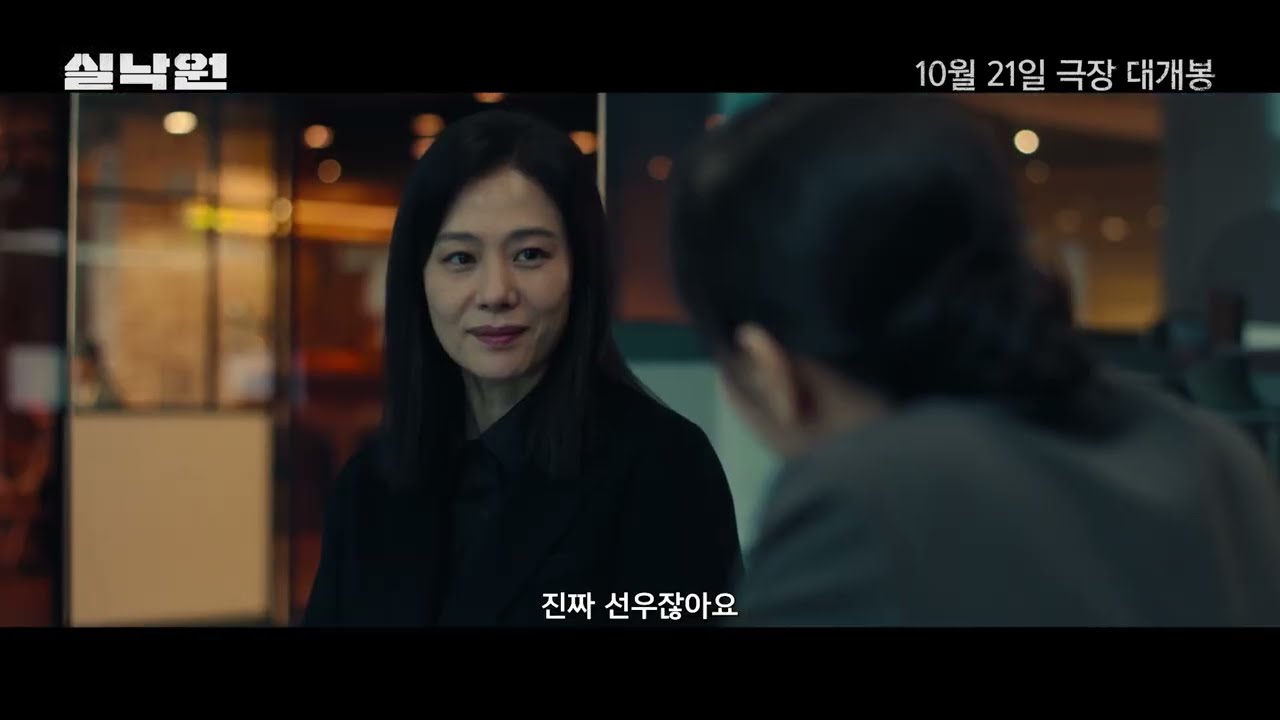 어두운 실내에서 정면을 보는 검은 옷 여성과 하단 자막