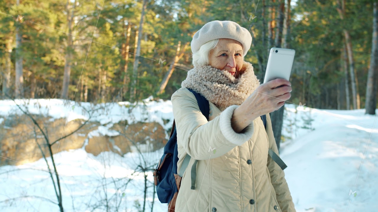 Happy elderly lady taking selfie with smartphone camera posing in park in winter