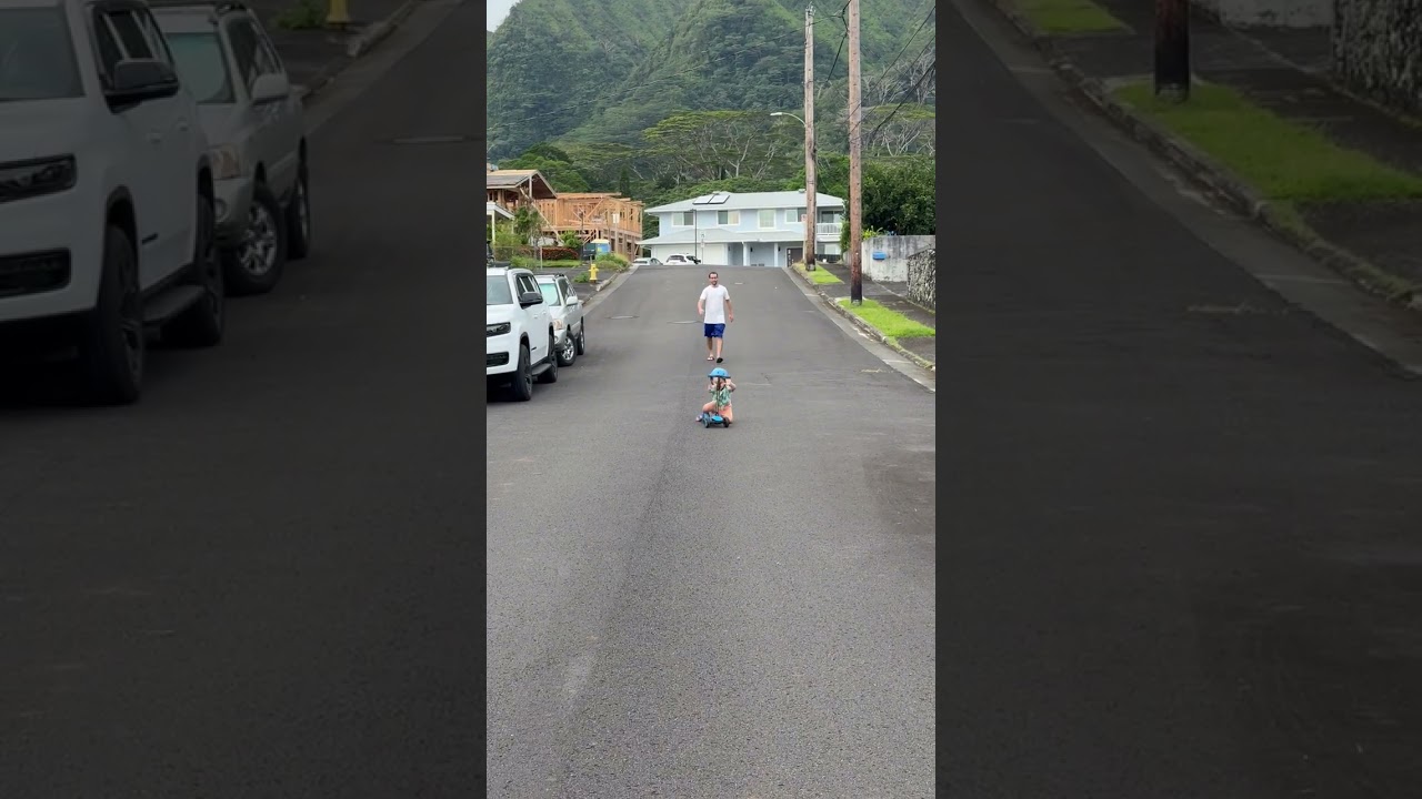 Boy in blue helmet rides scooter downhill on street then speed wobbles and faceplants as dad walks