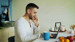 Handsome Young Man Talking Phone And Using Laptop Computer Sitting In The Kitchen After Breakfast In