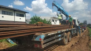Crane truck lifts steel sheet pile with hydraulic power in preparation for pile driving.