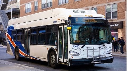 A New Flyer XN40 transit bus Driving  on an urban street with a brick building in the background.