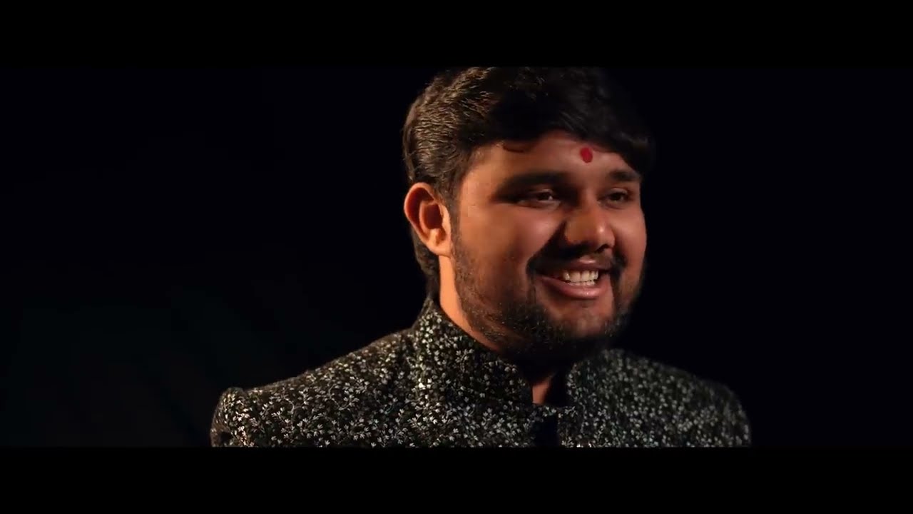 Uday Dhadhal smiling against a dark studio background in a sequinned black jacket — still from the Meldi Vare Chade music video