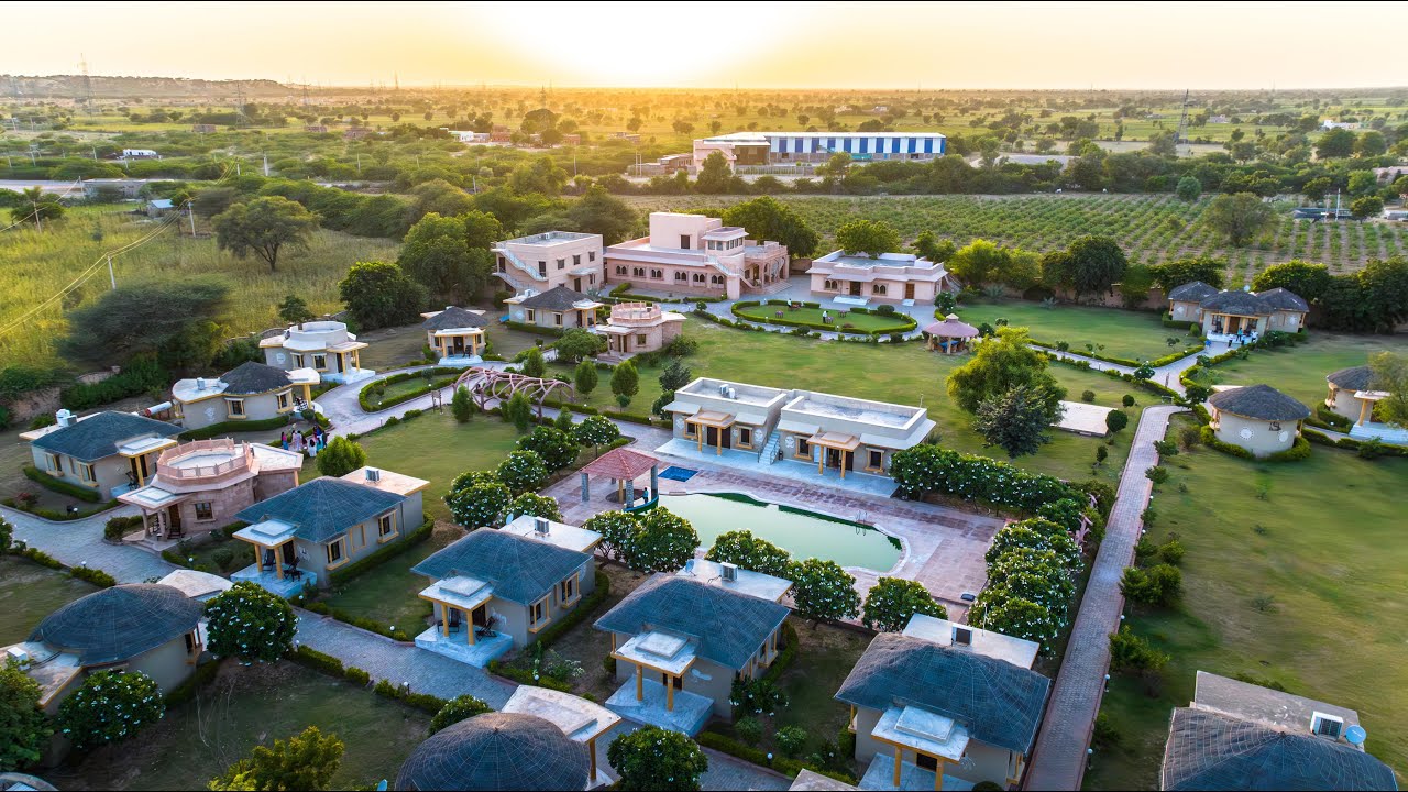 Aerial view of a resort at sunset with lush gardens, pathways, round huts, and a central pool, surrounded by green fields and scenic landscape.