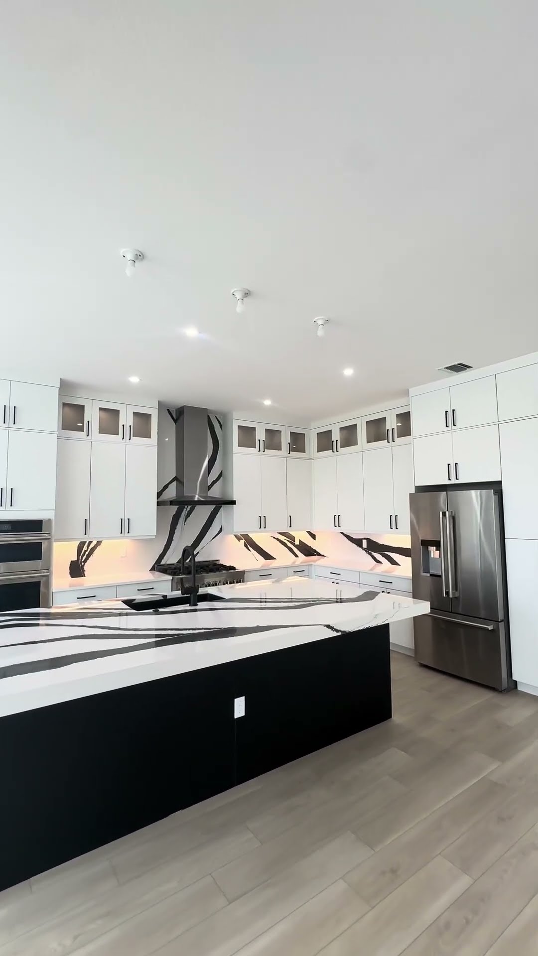 Modern kitchen with white and black cabinetry, a central island featuring a built-in stove and black sink. Sleek black and white marble backsplash extends to upper cabinets. Stainless steel refrigerator, wooden floor, and recessed lighting complete the space.