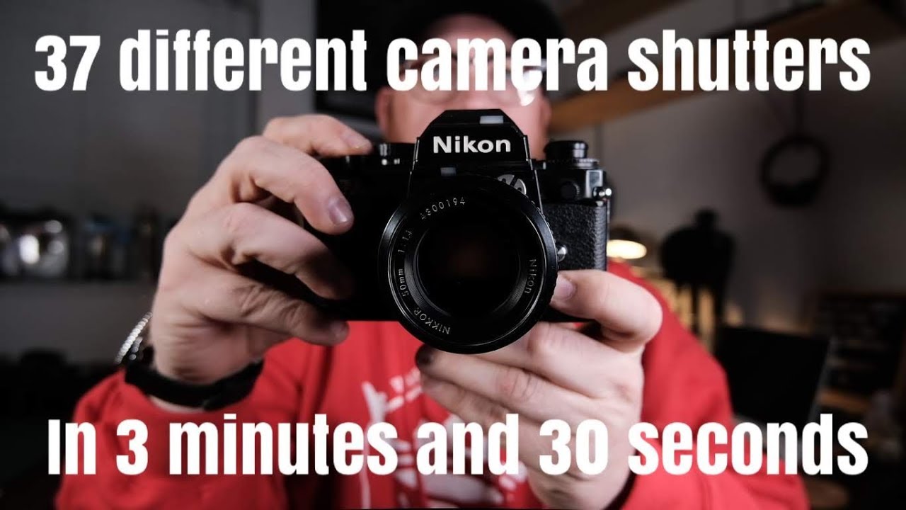  A man in a red shirt is holding a black and silver camera. The camera has a black lens. The man is holding the camera in his right hand. The man is looking at the camera. The camera has a setting to disable the shutter sound.