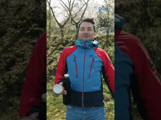 Мъж в цветно яке на открито, държи бутилка и се усмихва, наслаждавайки се на природата.